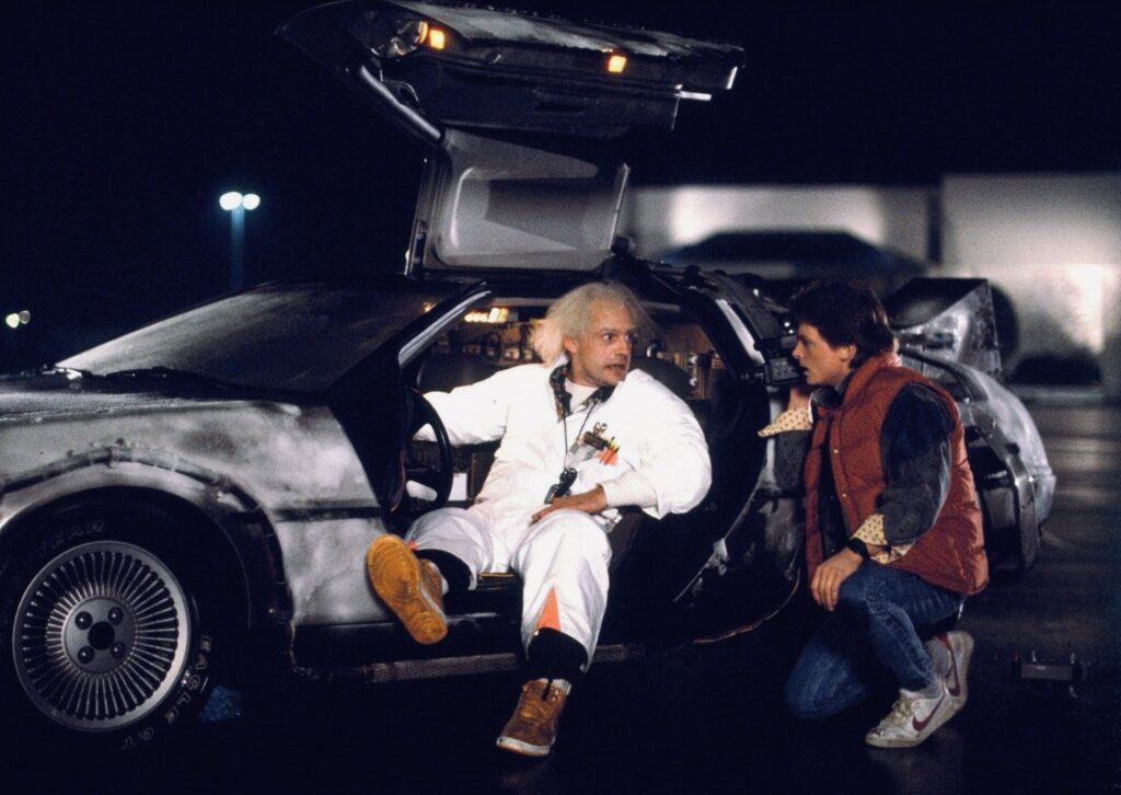 escena regreso al futuro 1985 Escena icónica de la película ‘Regreso al Futuro’ (1985) donde el Dr. Emmett Brown, con su característica bata blanca y expresión entusiasta, está sentado en el DeLorean, el automóvil convertido en máquina del tiempo. Frente a él, Marty McFly, vestido con su chaleco rojo, lo observa con asombro en un estacionamiento nocturno iluminado por farolas.